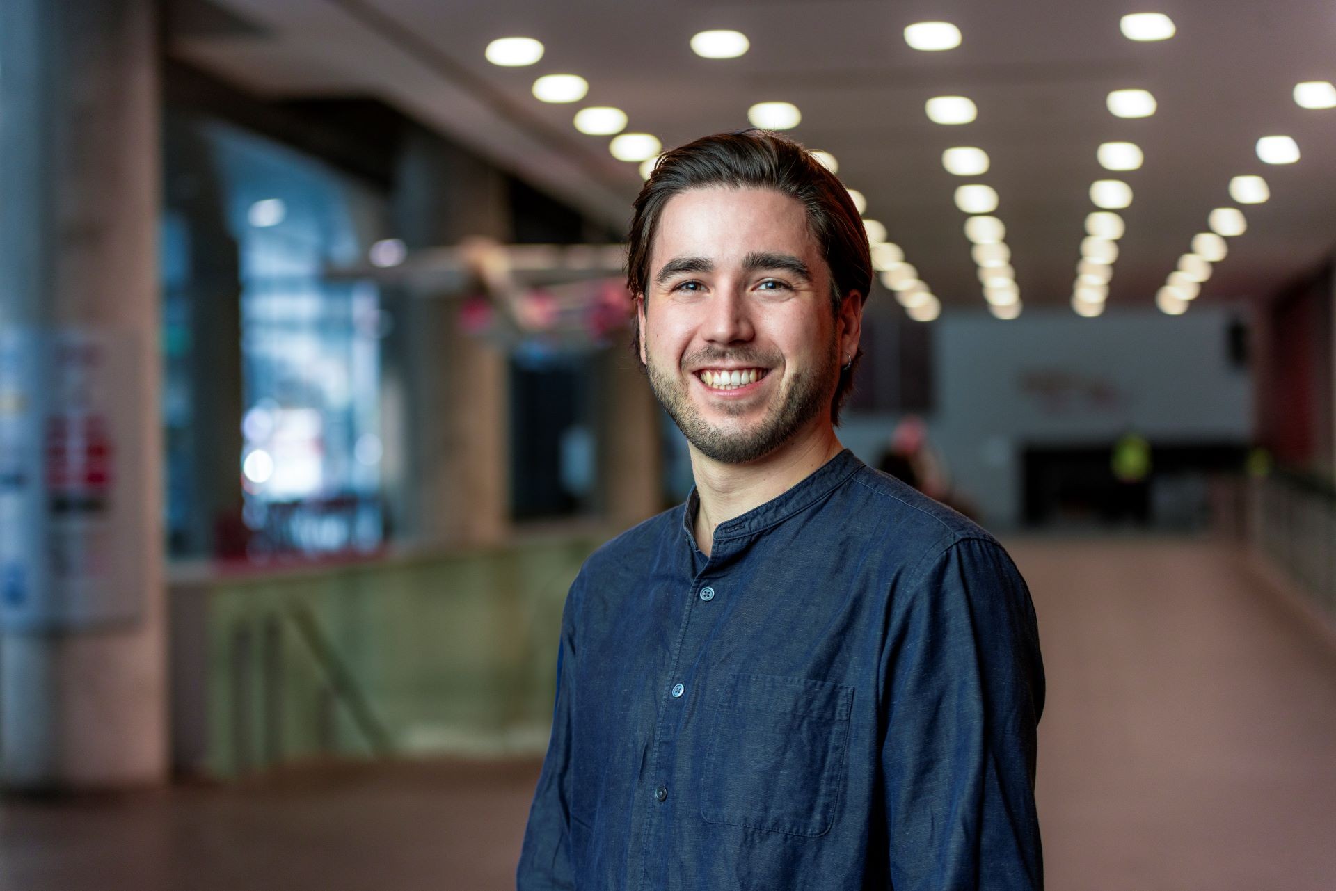 A young man in a blue shirt smiles in a university corridor