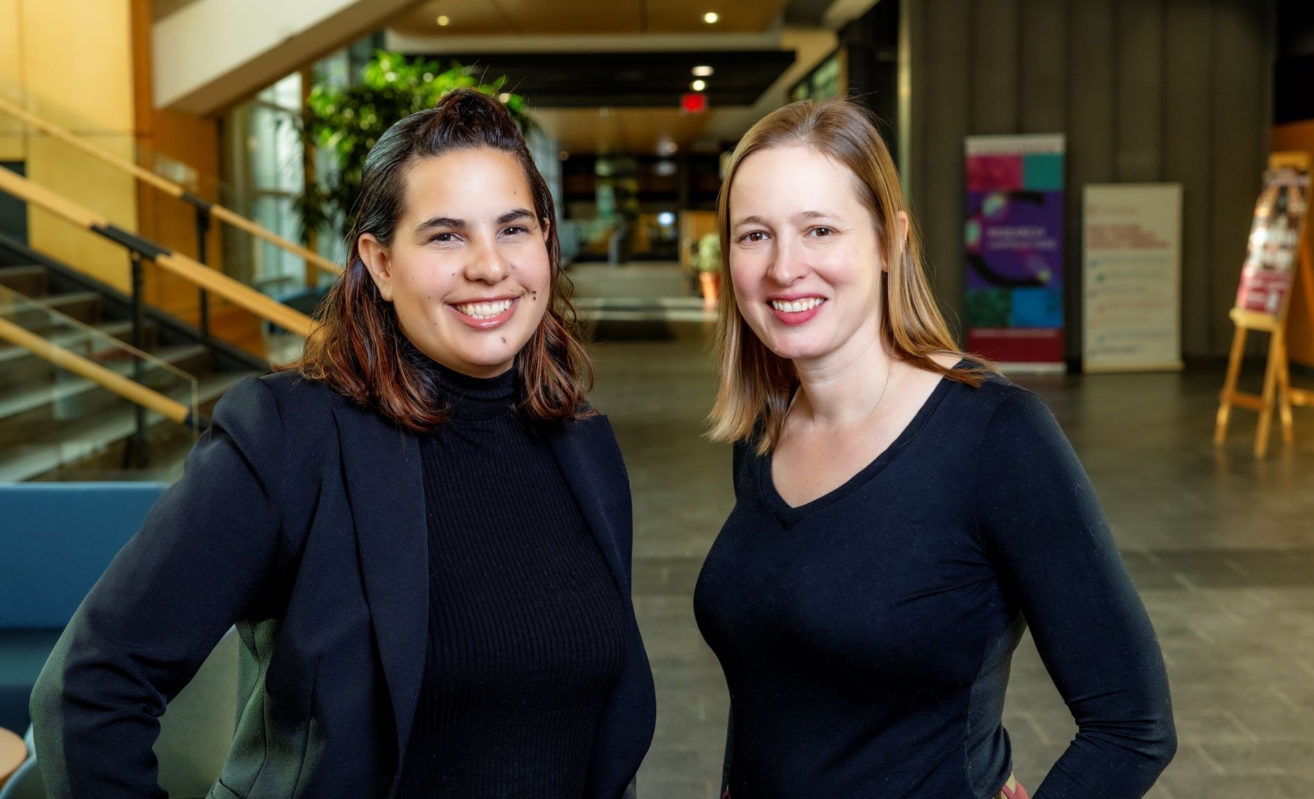 Two women dressed in dark clothes smile in a university corridor