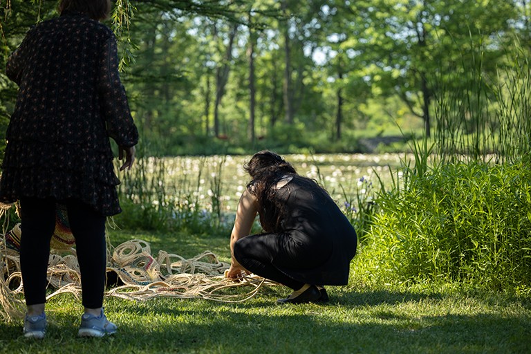 Two women on a grassy bank beside a river, with one crouching down to work on a rope artwork