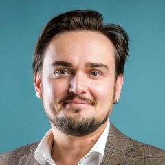 Homme souriant aux cheveux bruns courts, à la barbe et à la moustache courtes, portant une chemise à col blanc et une veste de costume bleue, sur fond turquoise.