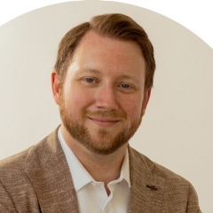 Homme caucasien souriant, cheveux courts roux, barbe courte et moustache, portant une chemise à col blanc et une veste de costume beige, sur fond beige.