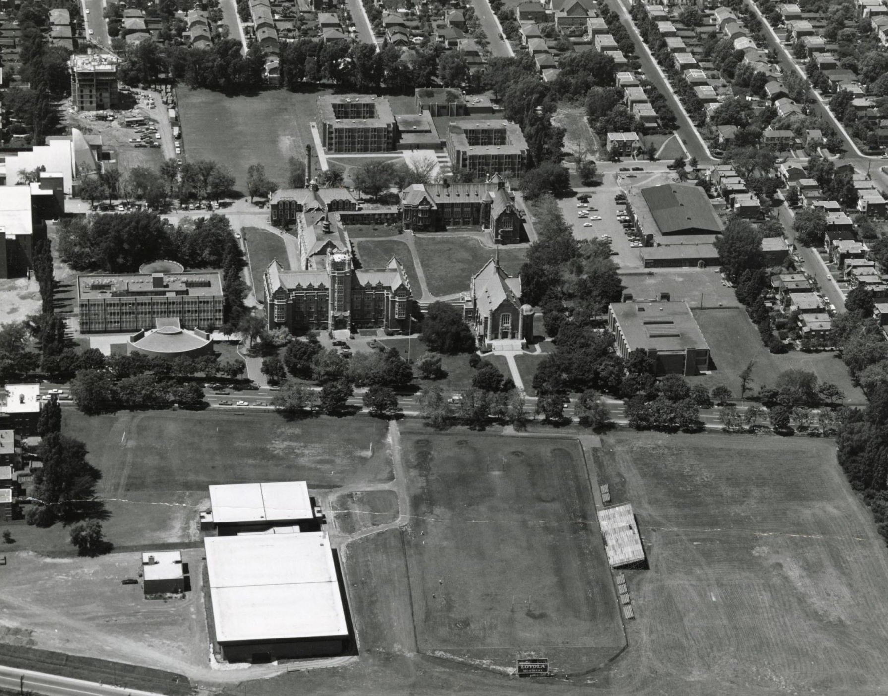 Photo aérienne en noir et blanc d'un campus comprenant plusieurs bâtiments et les rues résidentielles environnantes, montrant un vaste espace ouvert.