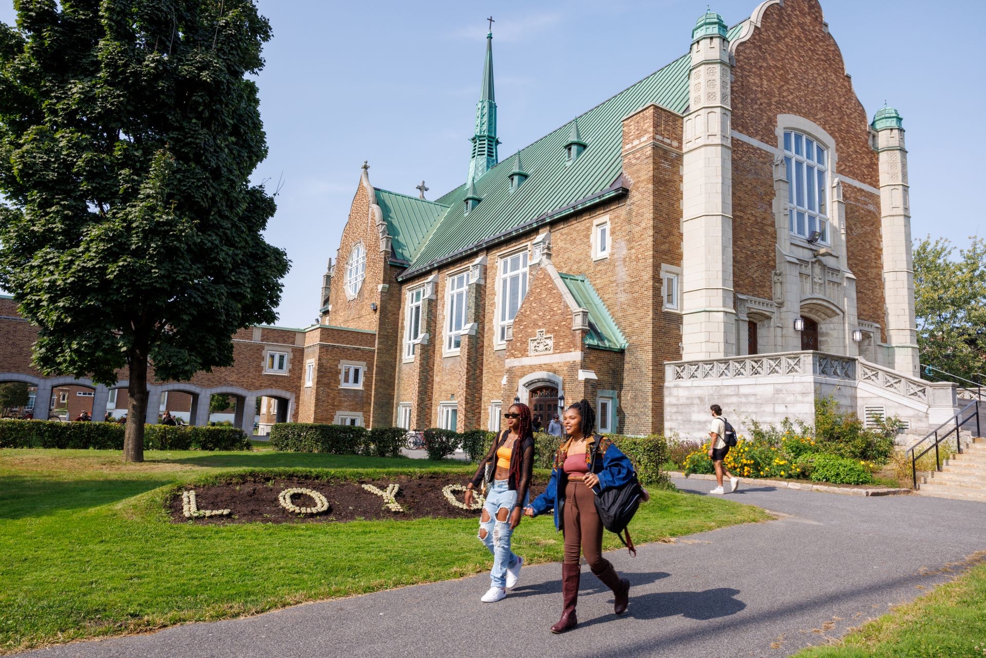 Deux étudiants passent devant le bâtiment historique du campus de Loyola avec, au premier plan, le mot « LOYOLA » écrit en toutes lettres dans un parterre de fleurs.
