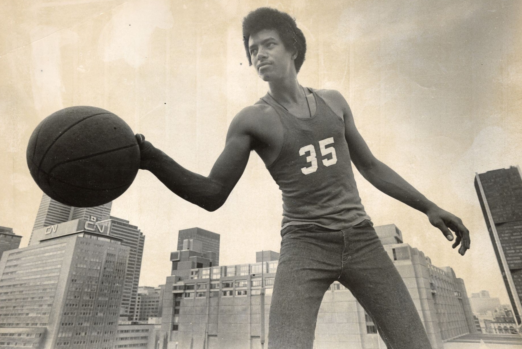 Une photo vintage en noir et blanc d'un jeune homme tenant un ballon de basket avec les gratte-ciel de la ville en arrière-plan, évoquant un sentiment de réussite sportive historique.