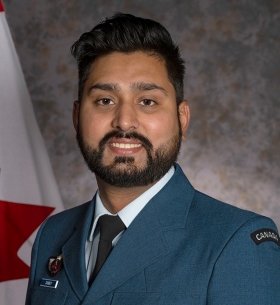 Un homme aux cheveux et à la barbe foncés sourit à la caméra. Il porte une veste bleue de l'armée de l'air