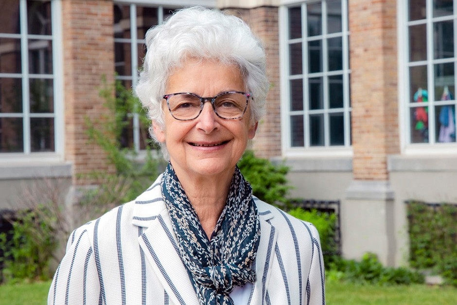 Corinne a des cheveux blancs, des lunettes et porte une veste blanche avec des rayures marines.