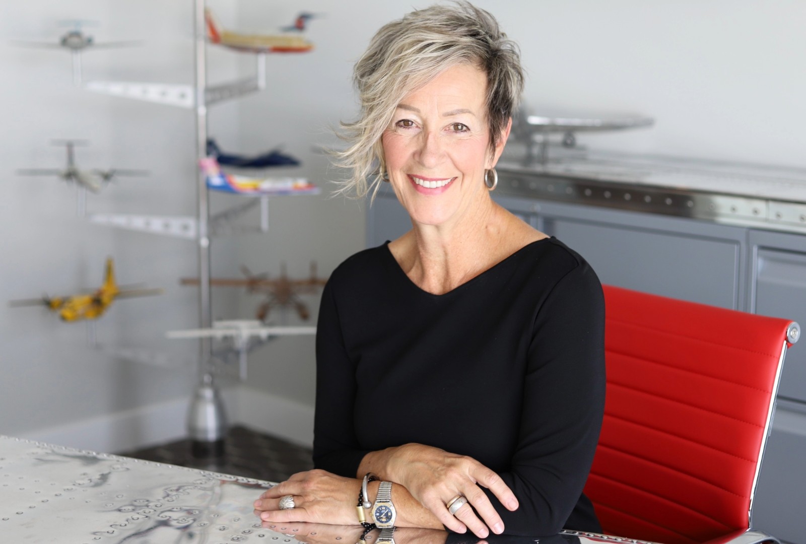 Une femme aux cheveux courts gris-blancs est assise sur une chaise rouge dans un bureau. Elle a les bras croisés à son bureau et porte un pull noir à col en V.