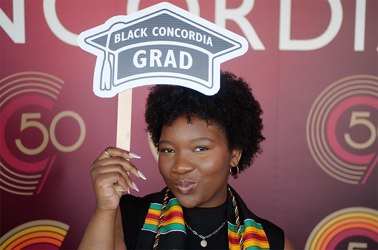 Jeune femme souriante aux cheveux noirs et bouclés, vêtue d'un haut noir et tenant une petite pancarte sur laquelle est écrit "Black Concordia grad".