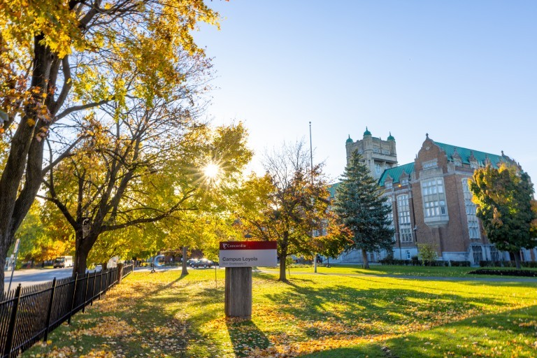 Un campus universitaire en automne, avec la lumière du soleil qui traverse les arbres. Un panneau indiquant « Campus Loyola » et un bâtiment en briques avec des toits verts sont visibles à l'arrière-plan.