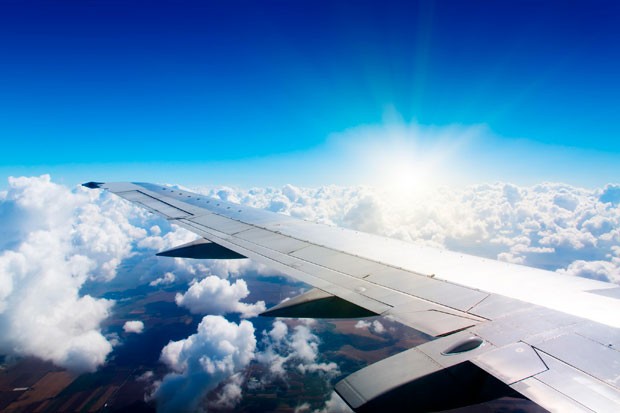 Vue depuis la fenêtre d'un avion montrant l'aile, avec un ciel bleu clair et le soleil brillant au-dessus d'une mer de nuages blancs et duveteux.