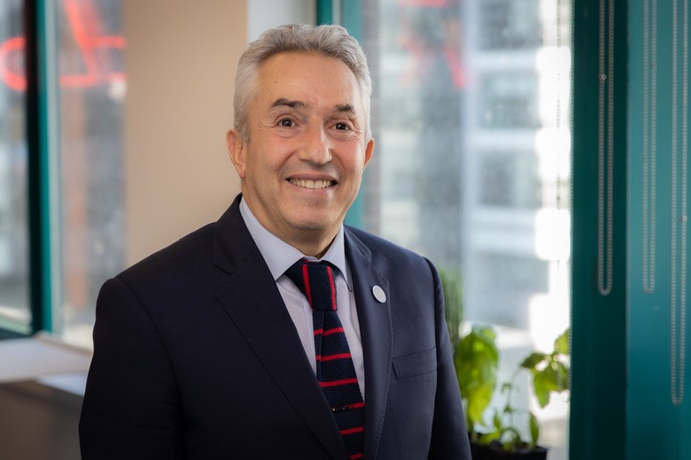 Portrait de Karim Zaghib souriant debout dans un environnement de bureau avec de grandes fenêtres. Il porte un costume sombre, une chemise bleu clair et une cravate rayée rouge et bleue. En arrière-plan, il y a des plantes vertes et une vue urbaine visible à travers les fenêtres.