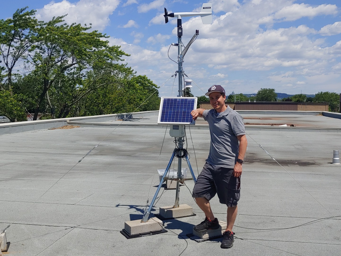 Student Daniel Baril standing on a rooftop next to weather monitoring equipment, including a solar panel, anemometer, and various sensors, against a backdrop of trees and a clear, partly cloudy sky.